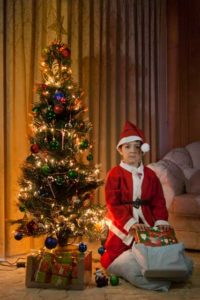 boy dressed up as santa claus opening presents with christmas tree lights